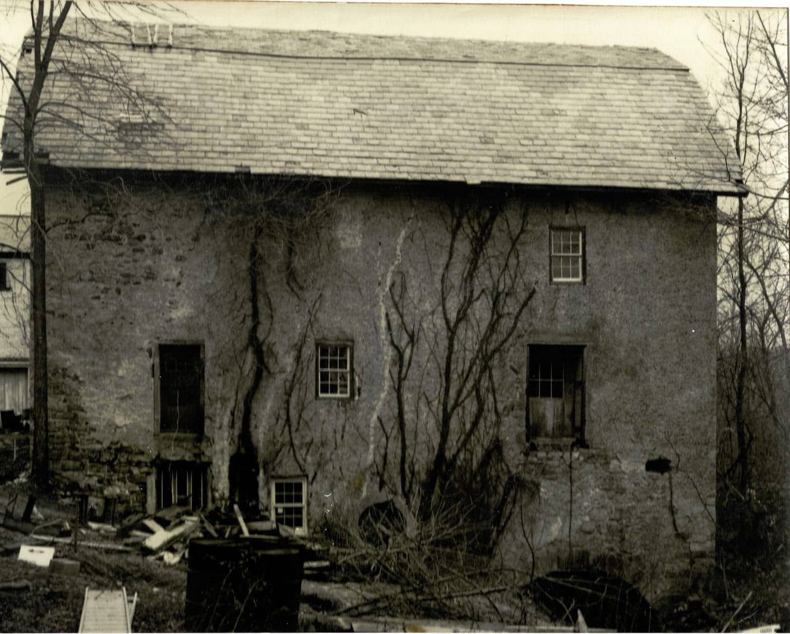 The mill before restoration, its stone walls covered in grey plaster, with bare winter trees and debris piled at the base