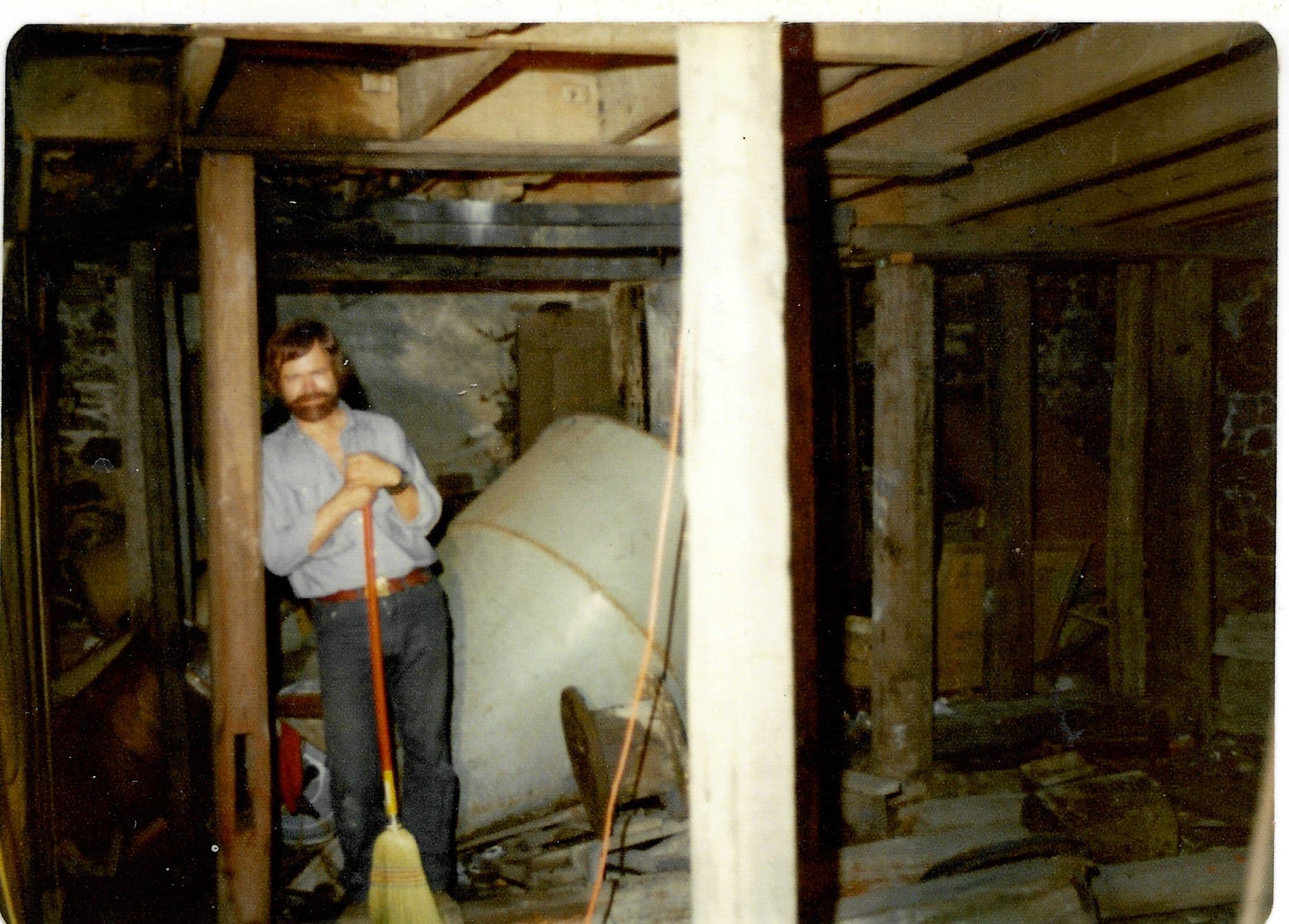 Charles Buckley standing with a broom in the gutted interior of the mill, beside a metal tank, surrounded by bare timber framing